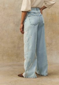 Jeans de pernera ancha en azul claro con bordado floral en el lado izquierdo, cinco bolsillos y una cintura alta. Fabricados en tela de mezclilla.