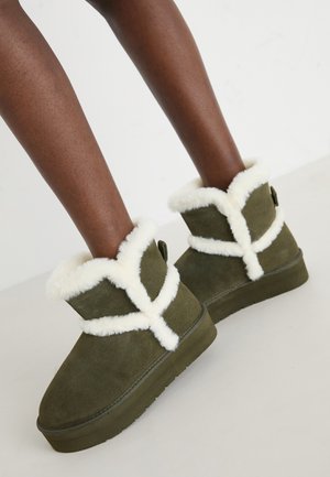 Botas de tobillo de ante verde oliva con un borde de piel sintética blanca y diseño de correas cruzadas, con una suela plana y superficie texturizada.