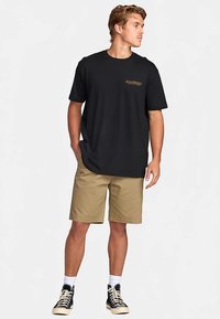 Svart t-shirt i bomull med korta ärmar, har en liten grafisk detalj i guld, kombinerad med khakifärgade shorts och svarta sneakers med högt skaft.