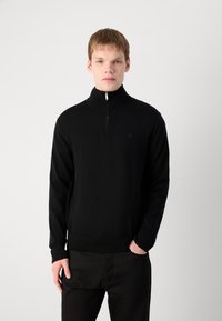 Melnas zip-augšdaļas puloveris, izgatavots no adīta auduma, ar garām piedurknēm, ribotām aprocēm un smalku logo uz krūškurvja. Apvienots ar tumšām biksēm.