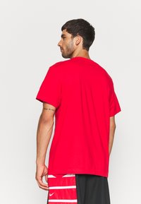 T-shirt en coton rouge à manches courtes et coupe ras du cou, vue de dos, associé à un short noir avec des rayures blanches verticales.