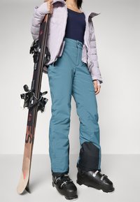 Ski oprema koja uključuje svijetlopurpuroljasti puf jaknu, tamnoplavi gornji dio, tirkizne hlače s crnim detaljima i crne skijaške čizme, držeći skije s vezovima.