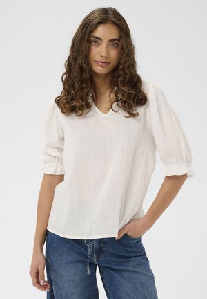 Femme aux cheveux bouclés portant un chemisier blanc à col en V avec des manches bouffantes et un jean bleu, debout avec une main dans la poche.