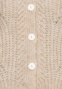 Beige stickat tyg med ett kabelmönster och tre jämnt fördelade runda vita knappar i en vertikal rad.