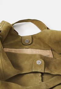 Olijfgroene suede tas met een zachte textuur aan de binnenkant, voorzien van een drukknoopsluiting, een crèmekleurige voering en een klein ritsvak.