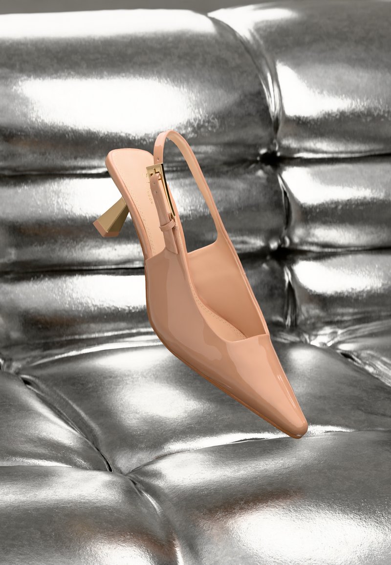 Lesklá nahá slingback vysoce podpatková bota se zlatou sponou a geometrickou patou odpočívající na prošívaném metalickém stříbrném povrchu.