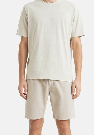 Uomo che indossa una maglietta beige a maniche corte e pantaloncini beige coordinati fino al ginocchio, in piedi davanti a uno sfondo bianco uniforme.