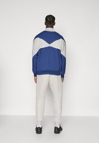 Blå og grå træningsdragt med en farveblok design; har en høj krave, elastiske manchetter og tapered bukser. Sorte sneakers fuldender looket.