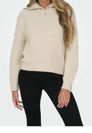 Vrouw draagt een beige gebreide trui met een kwartrits en ringtrekker, gecombineerd met zwart aansluitende broek, tegen een witte achtergrond.