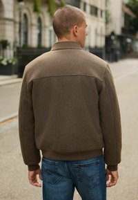 Jachetă din fleece maro cu guler înalt și manșete ribbate. Designul are o textură netedă și o croială lejeră, asociată cu blugi din denim albastru.