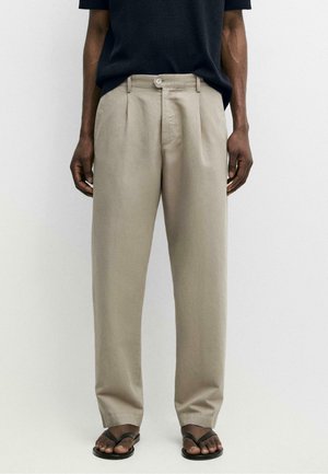 Mężczyzna ubrany w beżowe spodnie plisowane z zapięciem na guziki, czarny krótki top z krótkim rękawem i czarne sandały typu japonki, stojący na białym tle.