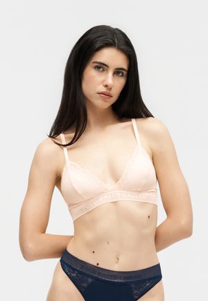 Kobieta o długich, ciemnych włosach, ubrana w bladoróżowy koronkowy biustonosz typu bralette i granatowe koronkowe majtki, stojąca na jednolitym jasnym tle.