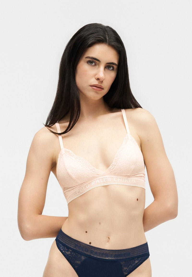 Kobieta o długich, ciemnych włosach, ubrana w bladoróżowy koronkowy biustonosz typu bralette i granatowe koronkowe majtki, stojąca na jednolitym jasnym tle.