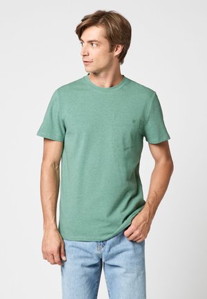 Homme portant un t-shirt vert à manches courtes avec une poche poitrine et un jean bleu clair, regardant vers sa gauche sur un fond uni.