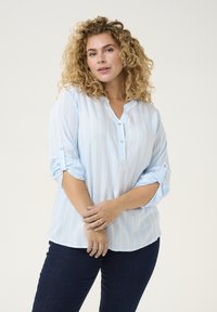 Jasnoniebieska koszula zapinana na guziki w pionowe białe paski, z podwiniętymi rękawami i luźnym krojem. Nosi ją z ciemnoniebieskimi dżinsami.