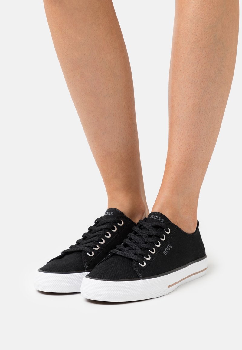 Personne portant des baskets basses noires avec semelles blanches et lacets noirs sur un fond clair, jambes visibles à partir du milieu du mollet vers le bas.
