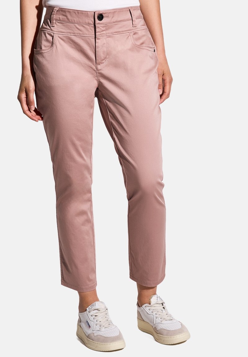 Personne portant un pantalon court rose clair et des baskets blanches, debout avec les mains détendues sur les côtés, sur un fond blanc.