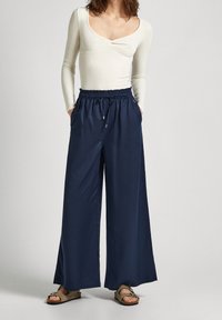 Kvinna klädd i en beige långärmad topp, lösa mörkblå vida byxor med dragsko i midjan och beige sandaler med spännen, stående mot en enfärgad bakgrund.