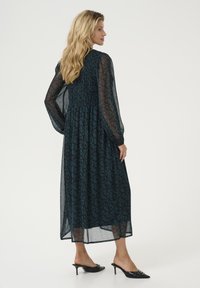 Long dark teal dress with sheer sleeves, featuring a floral pattern and smocked bodice. Paired with black heeled mules.