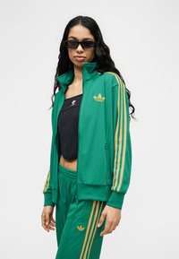 Γυναίκα που φορά πράσινο φόρμα Adidas με κίτρινες ρίγες, μαύρο τοπ που δεν φτάνει ως τη μέση και μαύρα ορθογώνια γυαλιά ηλίου, στέκεται μπροστά σε απλό φόντο.