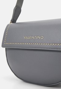 Šedá kožená crossbody taška s zaobleným tvarem, zlatými kovovými nýty a embosovaným logem na předním klapce.