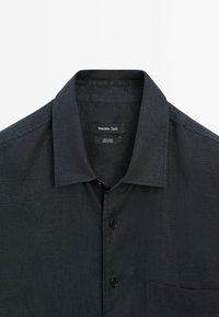 Camicia in lino grigio scuro con colletto classico, taschino singolo sul petto e bottoni neri. Tessuto strutturato con un sottile motivo a tessitura.