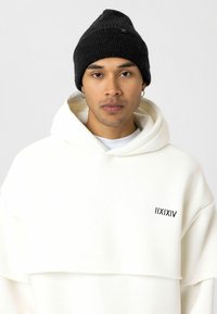 Witte oversized hoodie met een gelaagd ontwerp en zwart logo op de borst, gecombineerd met een gestructureerde zwarte gebreide beanie.