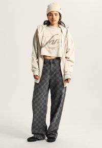 Beige, verkürzter Sweatshirt mit Logo, graue karierte Baggy-Hosen, Strickmütze und schwarze Slipper, die vor einem neutralen Hintergrund stehen.