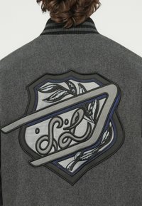 Osoba oblečená v sivej bunde s čiernym pruhovaným golierom a detailne vyšívanou nášivkou na chrbte so vzorom listov a abstraktných tvarov.