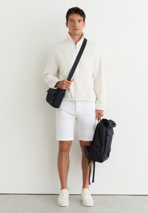 Hombre con jersey blanco con cremallera hasta la mitad, pantalones cortos blancos y zapatillas, sosteniendo una mochila negra en una mano y un bolso negro cruzado.