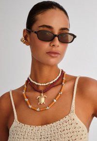 Γυναίκα που φορά στενά μαύρα γυαλιά ηλίου, πολλαπλά στρωμένα κολιέ μεταξύ των οποίων και μενταγιόν χρυσής χελώνας, χρυσά σκουλαρίκια-μανσέτες και μπεζ πλεκτό τοπ.