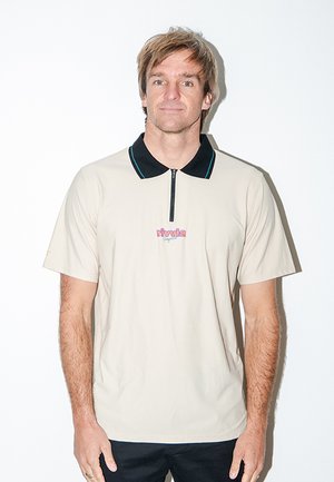 Mand med lys brunt hår iført en beige poloshirt med sort krave og lynlås, stående mod en hvid baggrund.