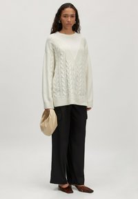 Chunky witte kabelgebreide trui met een V-hals, gecombineerd met zwarte wijde broek. Het model houdt een crèmekleurige clutch. Platte bruine schoenen.