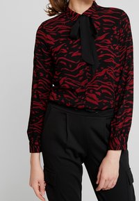 Blusa rossa e nera con motivo a strisce di tigre, maniche lunghe, chiusura frontale a bottoni e un dettaglio a farfalla nera al colletto.
