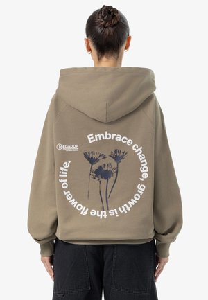 Person iført en brun hættetrøje med sort blomstergrafik og hvid tekst på ryggen, der lyder "Omfavne forandring, vækst er livets blomst".