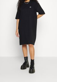 Černé tričkové šaty s krátkými rukávy, s malým vyšitým logem na hrudi a volným střihem. Spárováno s robustními černými šněrovacími botami.