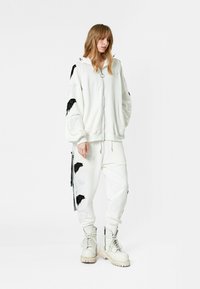 Sudadera blanca con cremallera y bordado de ala negra en las mangas, combinada con pantalones cónicos a juego con detalles similares. Botas blancas gruesas.