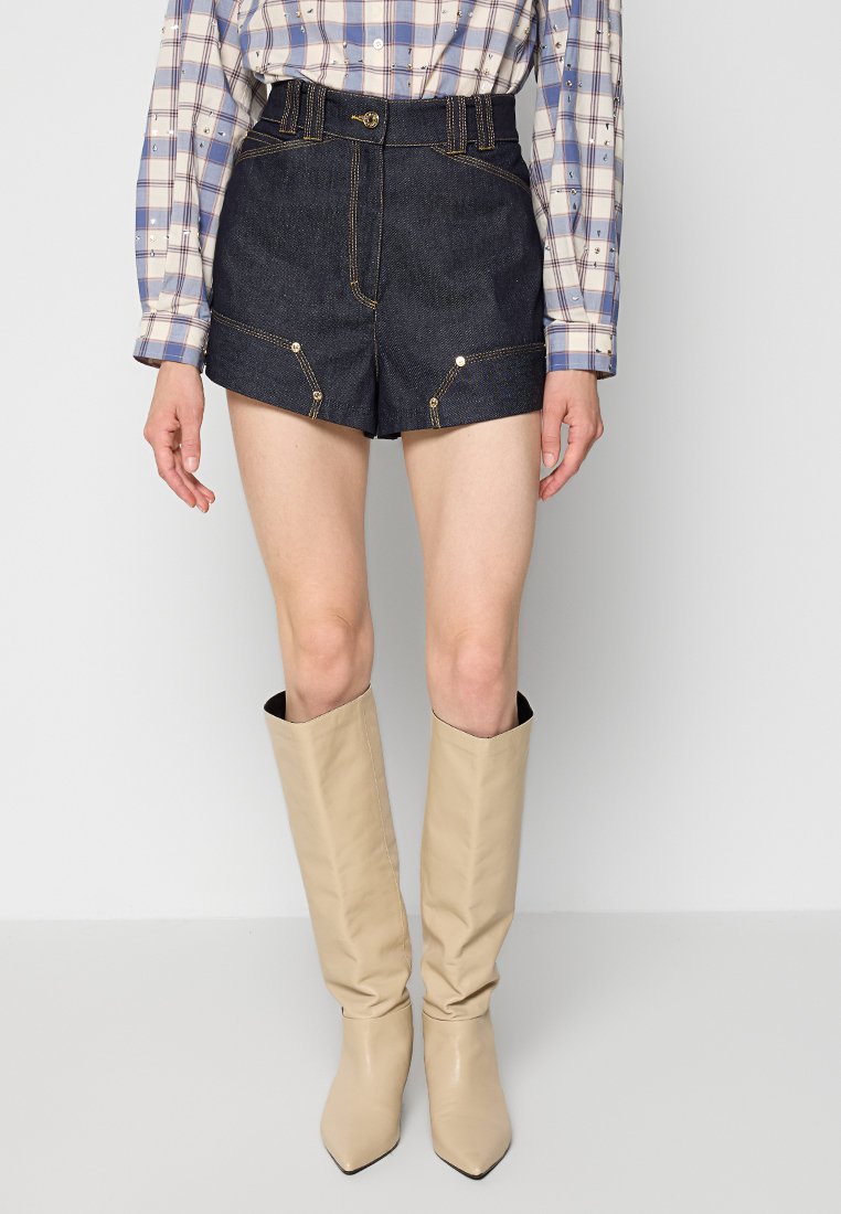 Denim-Shorts mit hoher Taille, goldenen Nähten und Vordertaschen. Kombiniert mit beigen kniehohen Stiefeln und einem karierten Button-Up-Hemd.