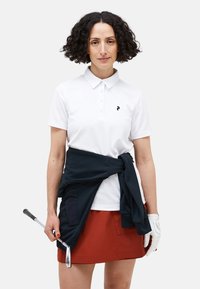 Witte poloshirt met een kraag en knopen, gecombineerd met een roestkleurige rok en een marineblauwe trui om de taille geknoopt. Golfclub in de hand.