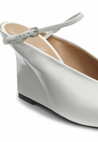 Chaussure compensée en cuir verni blanc avec bride à la cheville et boucle argentée, doublure intérieure beige, et semelle noire.