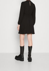 Mustapukuinen pitkähiha dressi, jossa on istuva vyötärö ja avainaukko selässä. Yhdistetty polven ylle ulottuviin mustiin nahkakenkiin, joissa on kengännauhat.