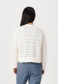Suéter blanco de punto con corte holgado, que presenta un patrón abierto texturizado. Dobladillo recortado y mangas largas, combinado con jeans de mezclilla azul.