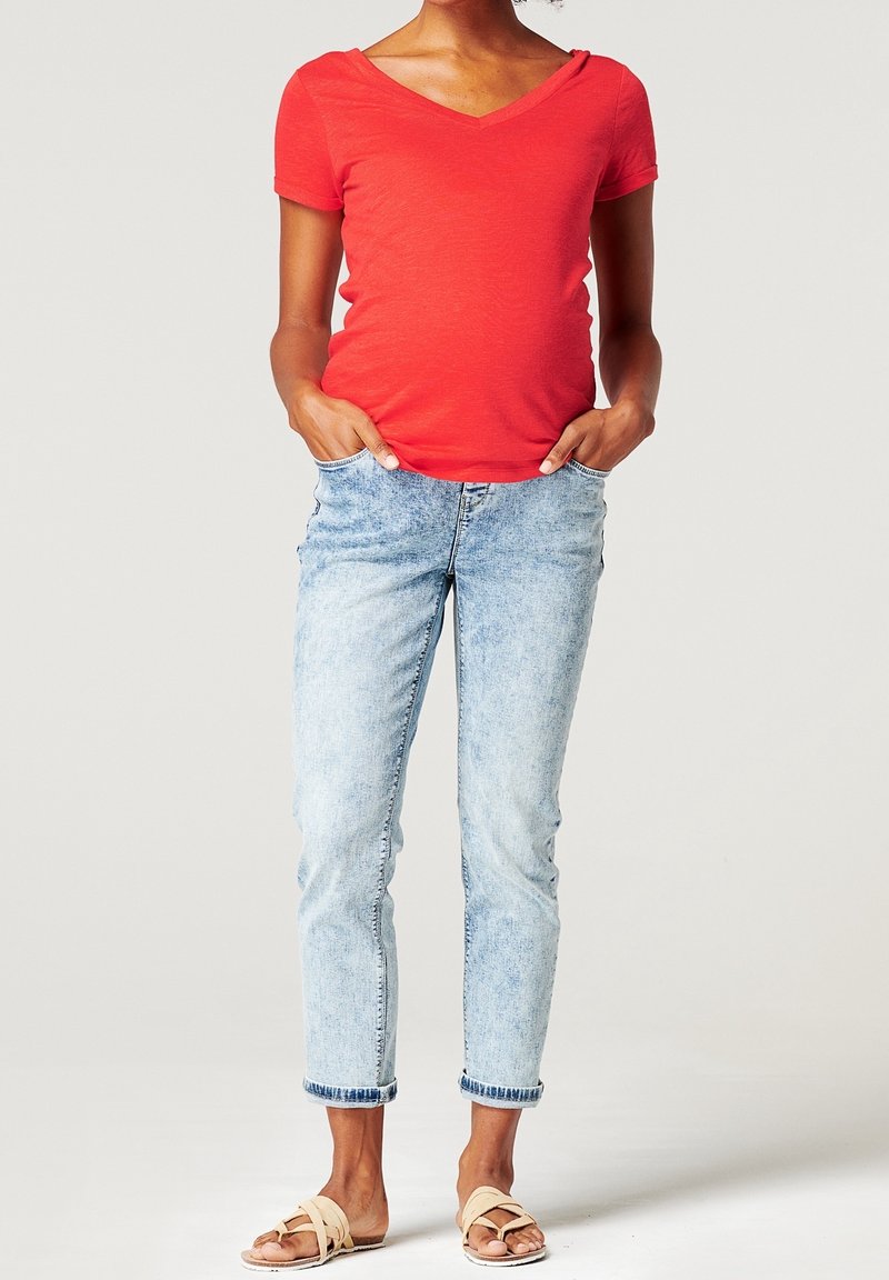 Personne portant un t-shirt rouge à col en V manches courtes, un jean bleu clair retroussé et des sandales beige à enfiler, debout les mains dans les poches.