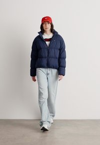 Granatowa kurtka puchowa z zamkiem z przodu, jasnoniebieskie dżinsy i czerwona czapka. Tkanina jest gładka, o pikowanej strukturze i elastycznych mankietach.