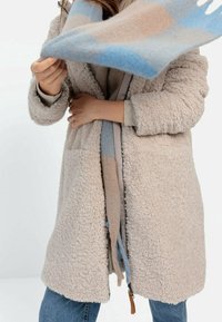 Manteau beige, duveteux et long avec une finition texturée et de grandes poches. Assorti à une écharpe multicolore avec des tons bleus, beigés et marron clair.