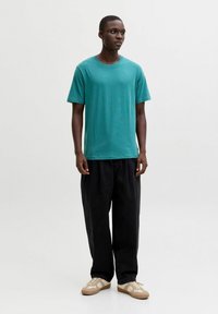 Homme debout portant une chemise à manches courtes couleur sarcelle, un pantalon noir ample et des baskets beiges, devant un fond blanc uni.
