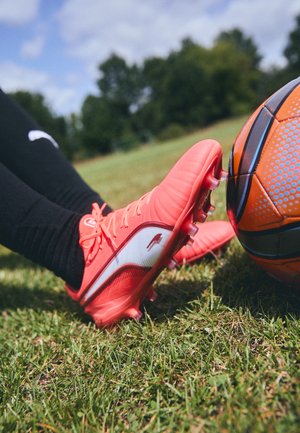 Scarpino da calcio rosa brillante con un accento bianco, superficie testurizzata e numerosi tacchetti, posizionato accanto a un pallone da calcio arancione su erba.