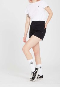 Short en denim noir avec revers roulés, associé à un t-shirt blanc et des chaussettes blanches avec un logo, porté avec des baskets noires. Texture lisse.