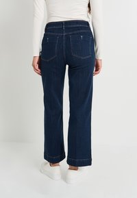 Personne portant un jean droit bleu foncé et des baskets blanches, se tenant contre un fond clair uni, vue de dos.