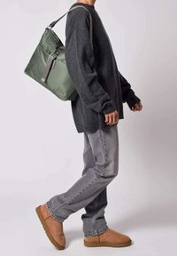Mochila de nailon verde con cierre superior y correa ajustable gris. Modelo que lleva puesta una camiseta de manga larga gris oscuro y jeans gris claro.